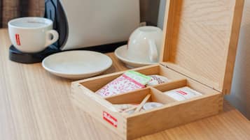 A wooden box with compartments containing tea bags and utensils, a white coffee maker, and white cups on a wooden table.