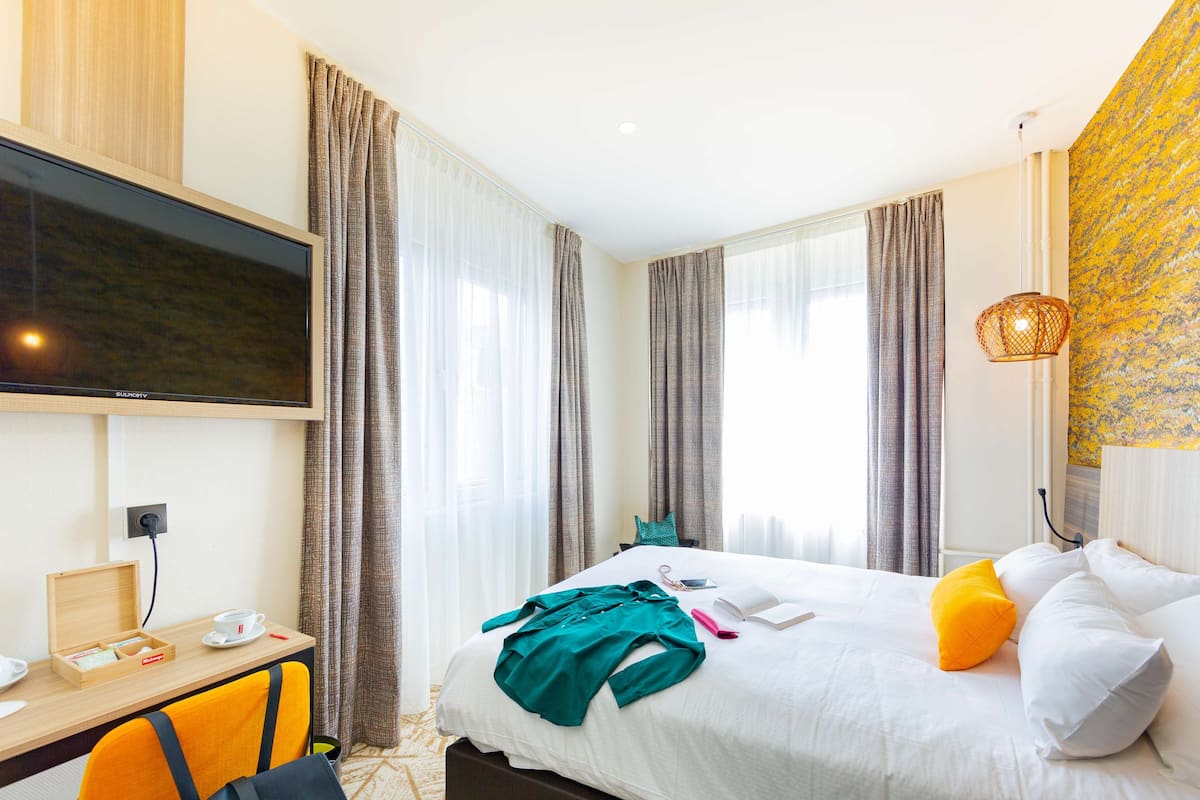 Una habitación de hotel con cama, escritorio con televisor, una silla y ventana con cortinas.