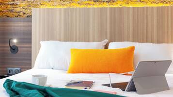 Una cama bien hecha con almohadas blancas y anaranjadas, una computadora portátil, una taza y un cuaderno sobre la cama.