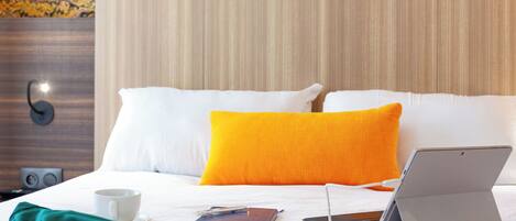 A neatly made bed with white and orange pillows, a laptop, a cup, and a notebook on the bed.