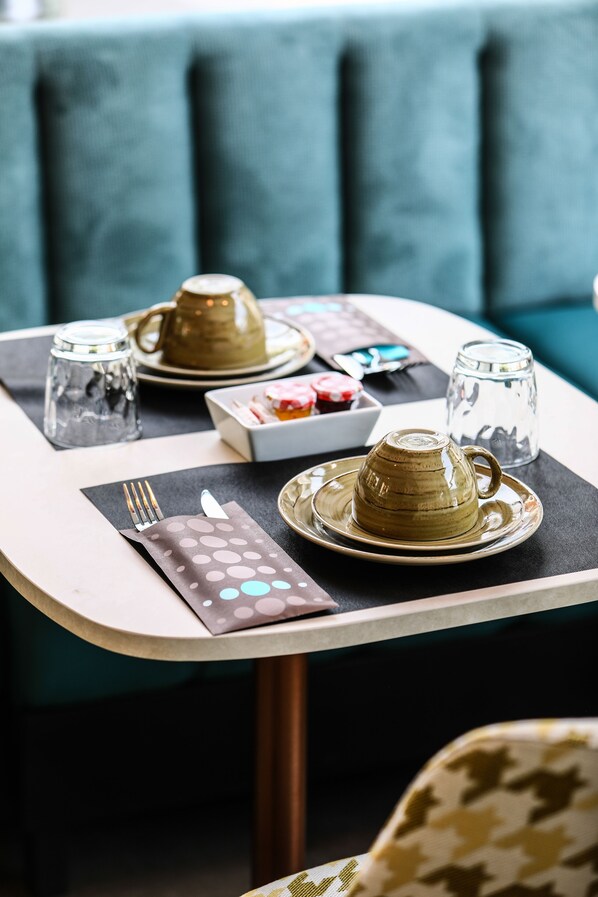 Una mesa dispuesta con un calentador de té, tazas y un plato pequeño de pasteles, un mantel individual estampado y un tenedor sobre un camino de mesa.