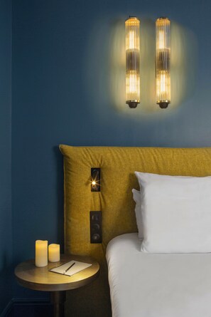 Quarto de hotel com cama, mesa de cabeceira redonda com velas e iluminação na parede.