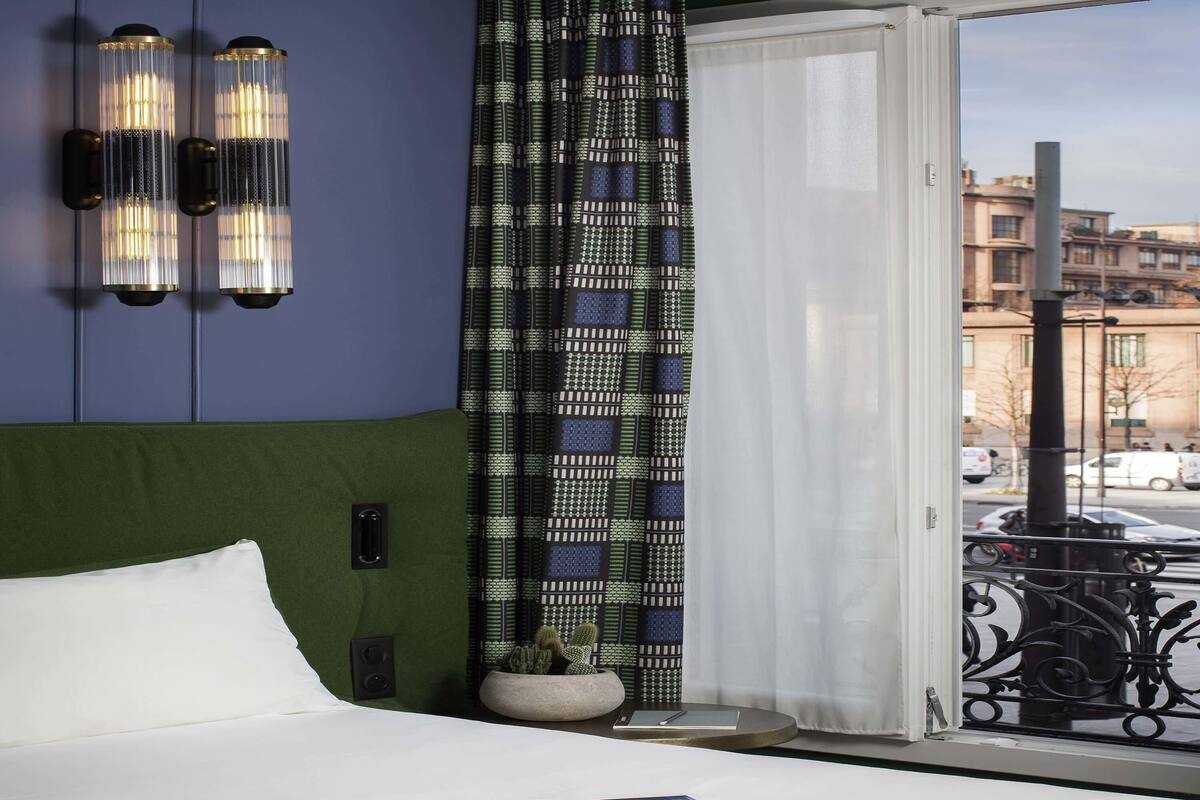 A hotel room with a bed, a window with a view of buildings, a patterned curtain, and wall sconces.