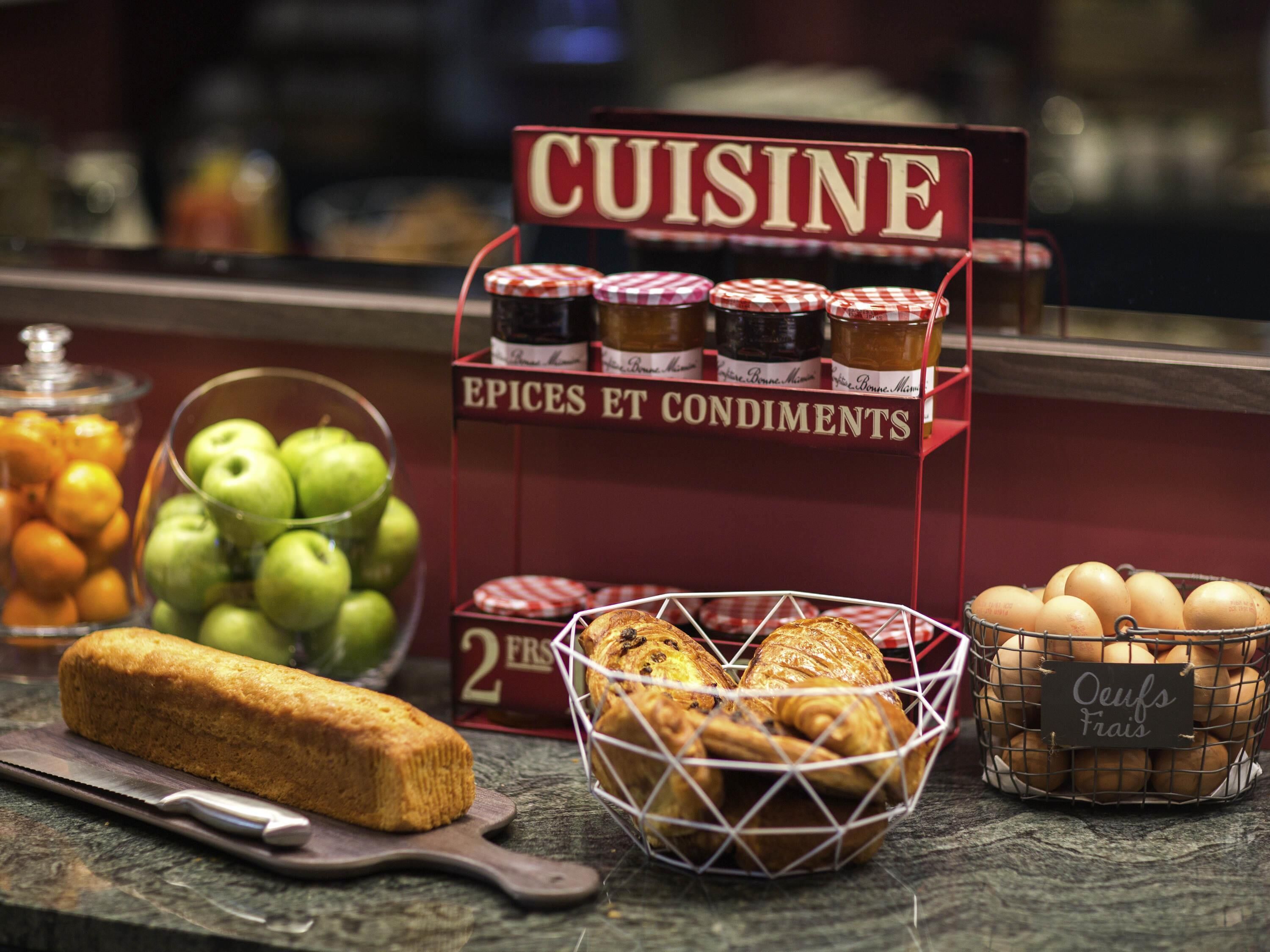 A display of food items including a loaf of bread, apples, jars of condiments, and a basket of eggs.