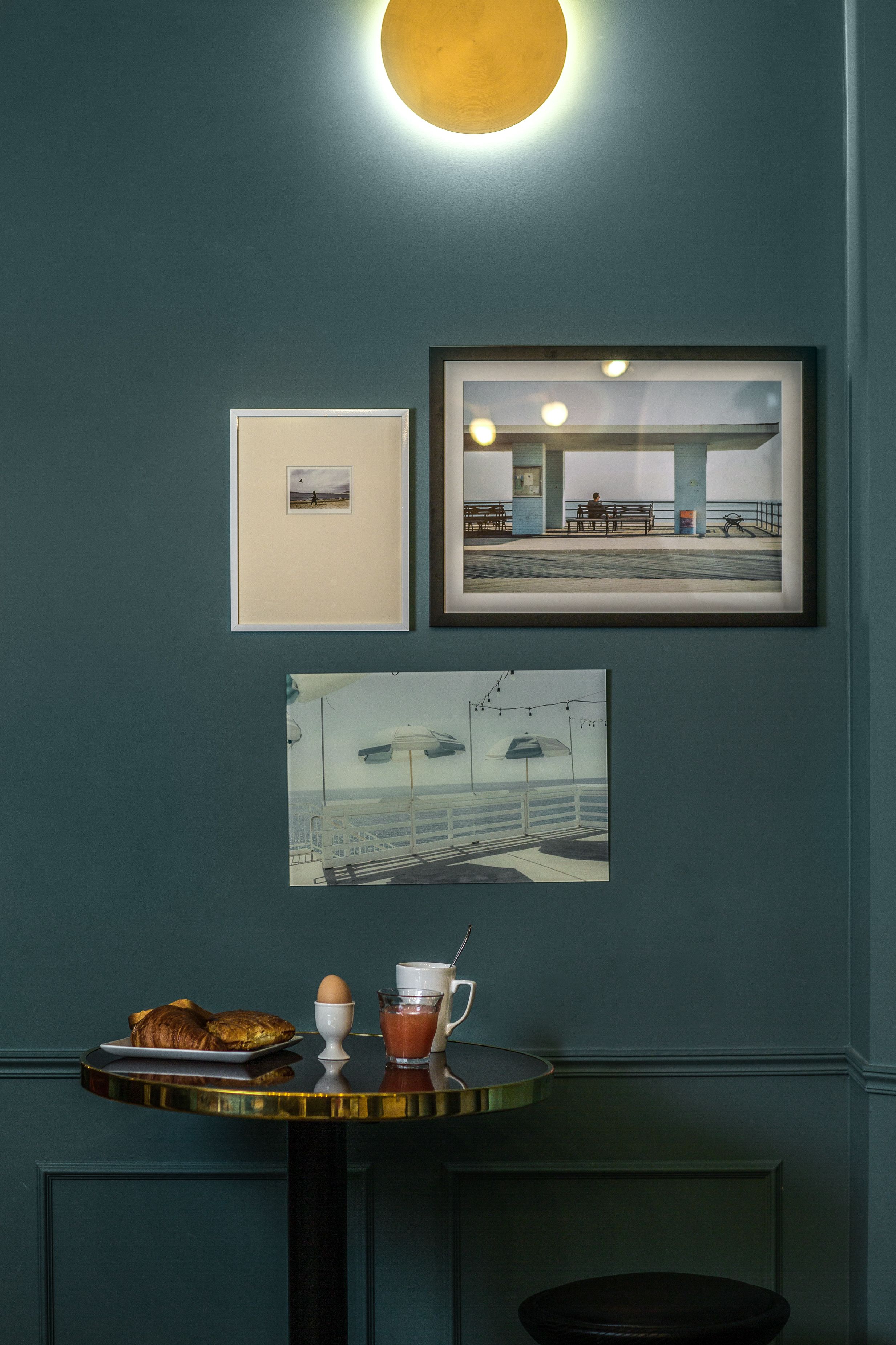 A wall with three framed pictures, a table with breakfast items, and a round table with a stool.
