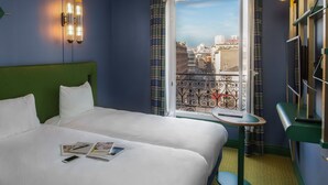 A hotel room with a large bed, a small table, a bookshelf, and a window with a city view. - ibis Styles Paris Nation Cours de Vincennes (Paris)