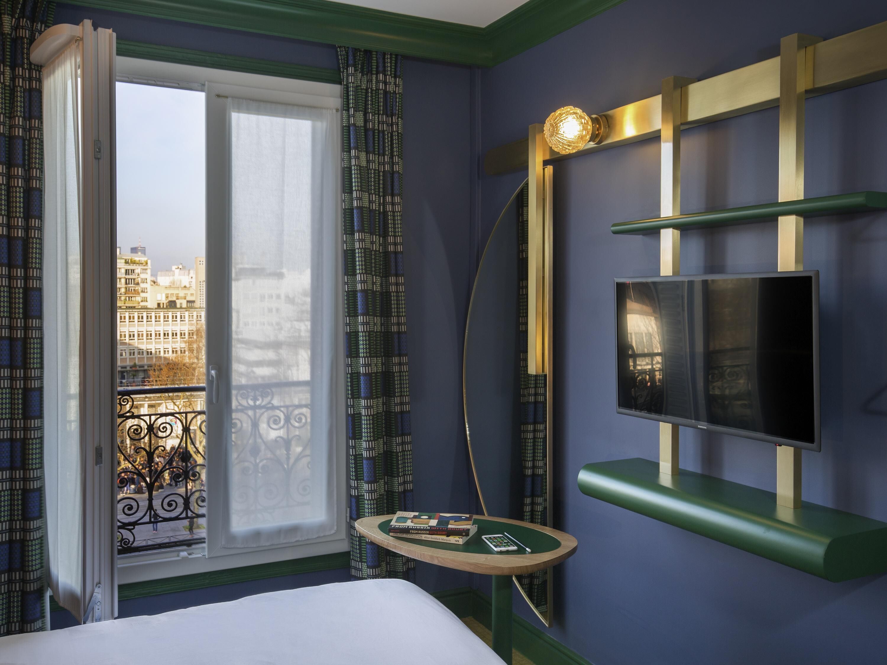 Habitación de hotel con cama, televisor montado en la pared, una mesita con libros y un control remoto, y balcón con cortinas estampadas.