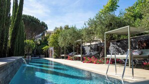Una piscina con pérgola y sillones de descanso, rodeada de árboles y flores.