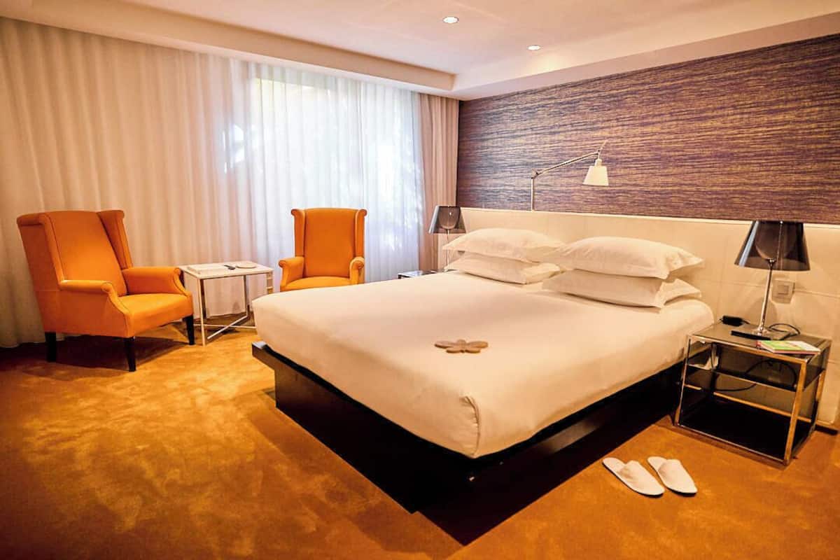 Een hotelkamer met een groot bed, twee oranje fauteuils, een nachtkastje, een lamp en een paar slippers op de vloer.