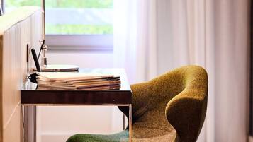 A desk with a stack of books and a lamp, a green chair, and a window with curtains.