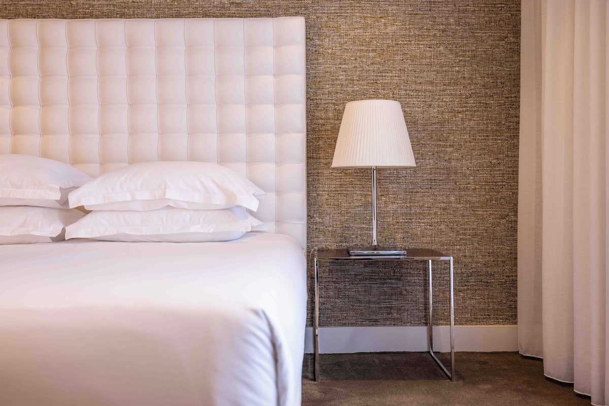 Een net opgemaakt bed met witte lakens en een geborduurde hoofdbord, een moderne lamp op een klein tafeltje ernaast, en een gestructureerde wand op de achtergrond.