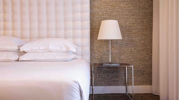 Una cama impecablemente vestida con sábanas blancas y un cabecero acolchado, una lámpara moderna sobre una mesita de noche pequeña, y una pared con textura al fondo.
