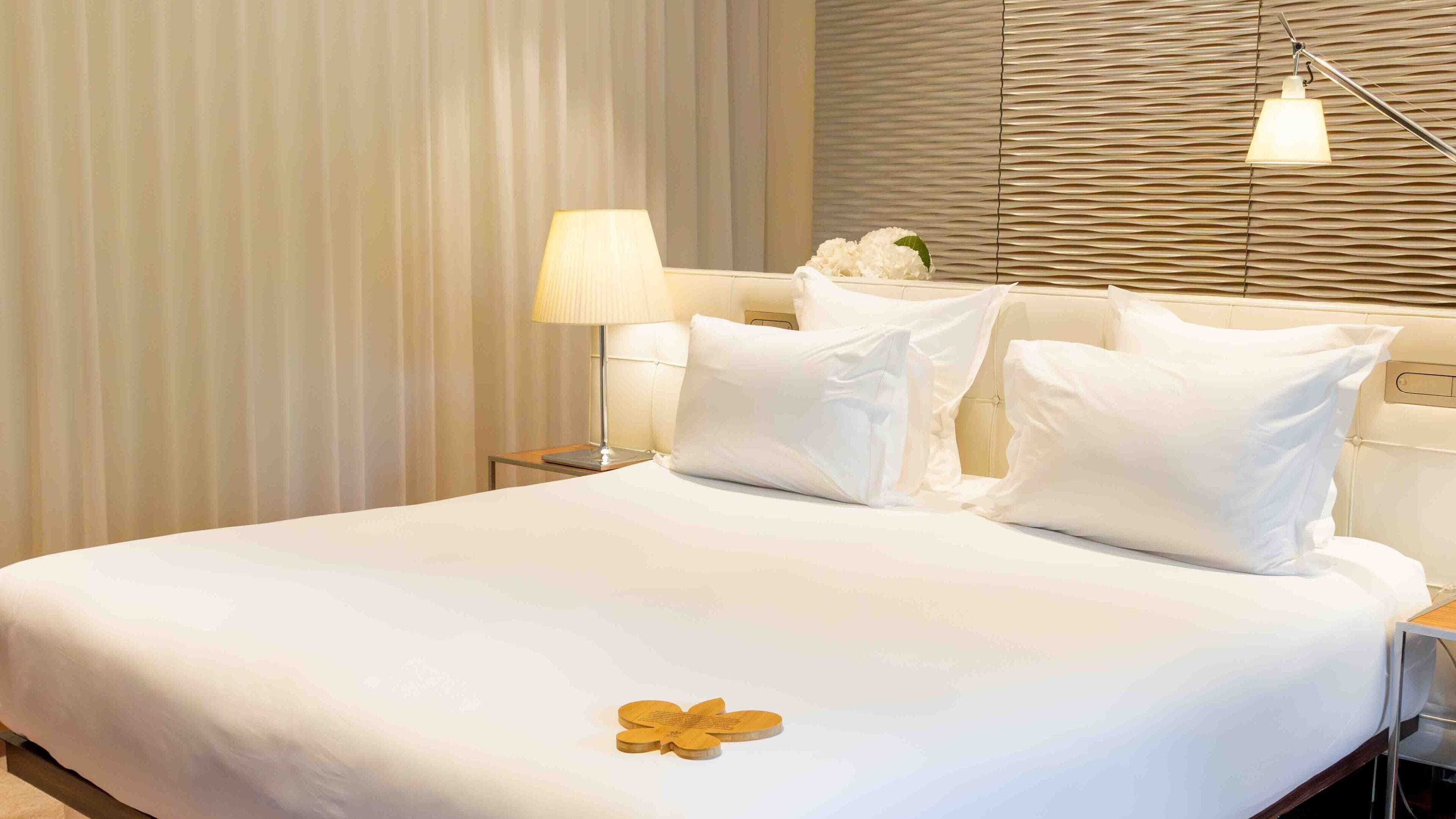 Habitación de hotel con una cama grande, ropa blanca, una lámpara de noche y una flor decorativa sobre la cama.