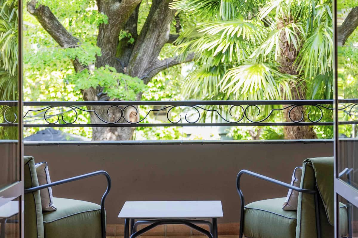 Een balkon met twee stoelen en een kleine tafel, met uitzicht op een weelderige groene tuin met bomen en planten.