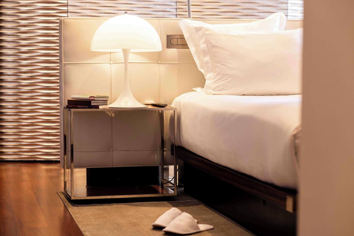 Een net opgemaakt bed met witte beddengoed, een bedlampje en een klein stapeltje boeken op het nachtkastje.