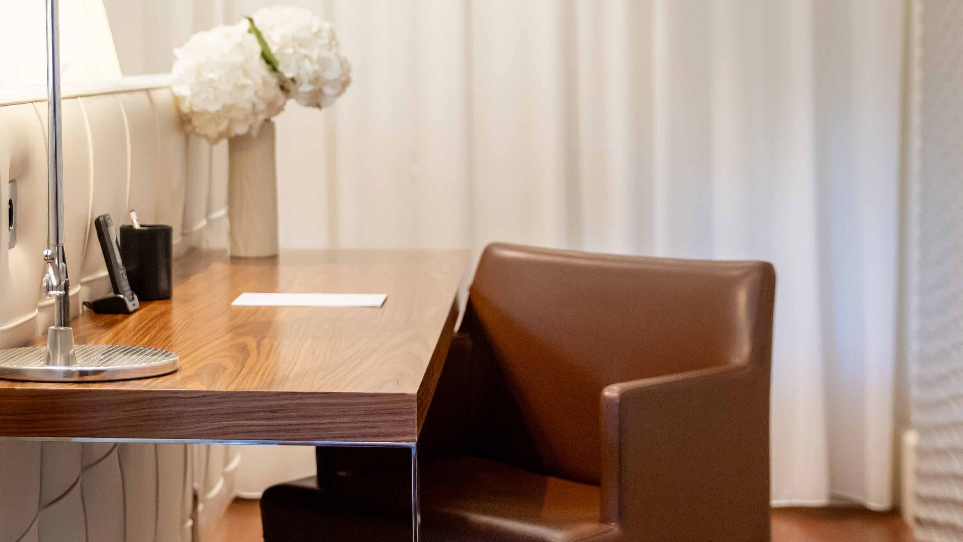 A hotel room with a wooden desk, a brown leather chair, a white vase with flowers, and a lamp.