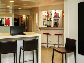 Un coin bar avec un ordinateur portable posé sur le comptoir, deux tabourets de bar et un réfrigérateur rempli de bouteilles.