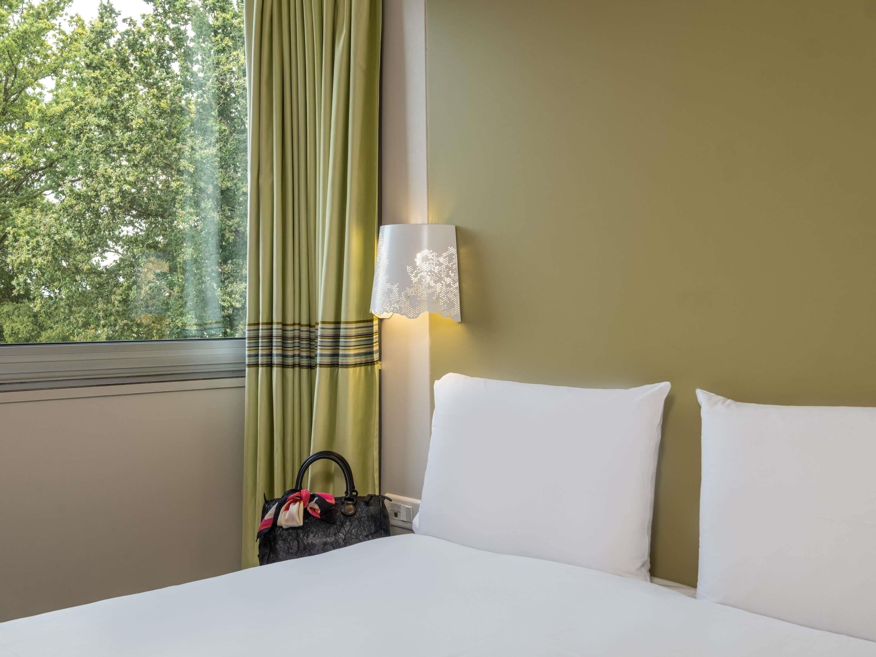 A hotel room with a bed, a lamp, a window with curtains, and a handbag on the floor.