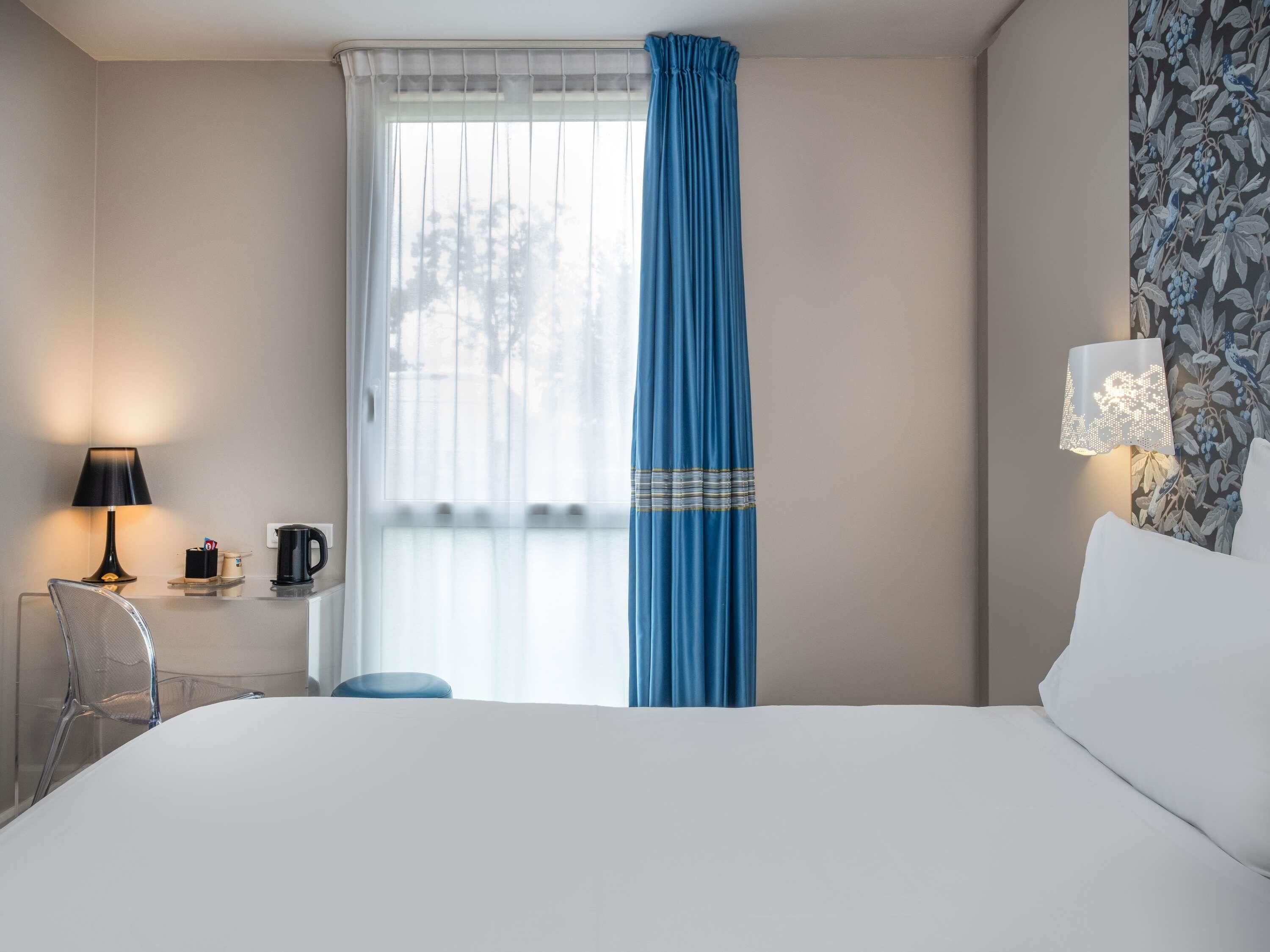 A hotel room with a bed, a desk with a lamp, a chair, a window with a blue curtain, and a wall with a floral pattern.