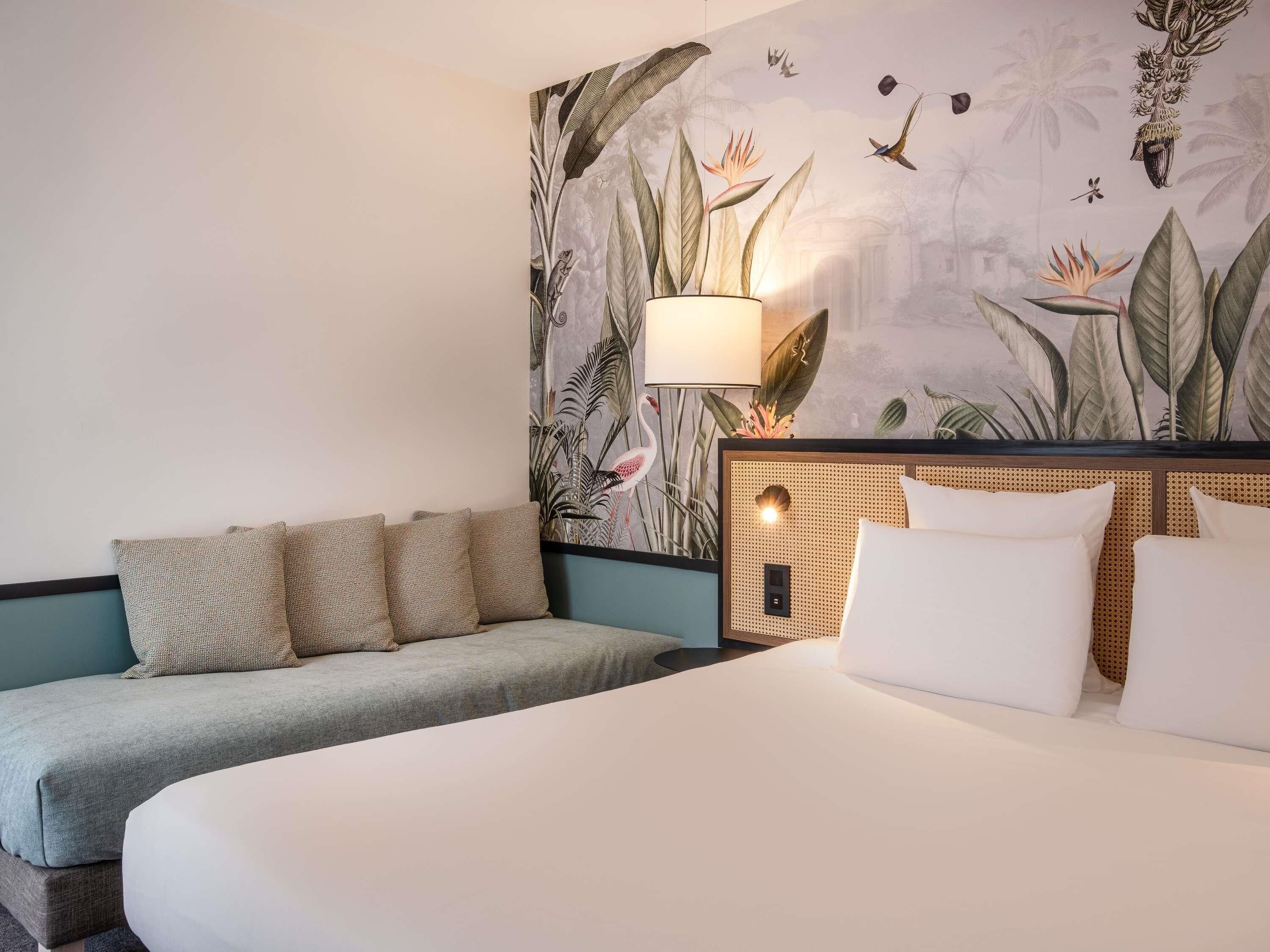 A hotel room with a bed, a sofa, a wall mural, and a lamp.