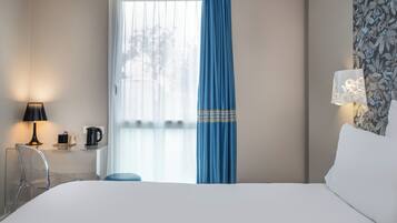 Habitación de hotel con cama, escritorio con lámpara, silla y ventana con cortina azul.