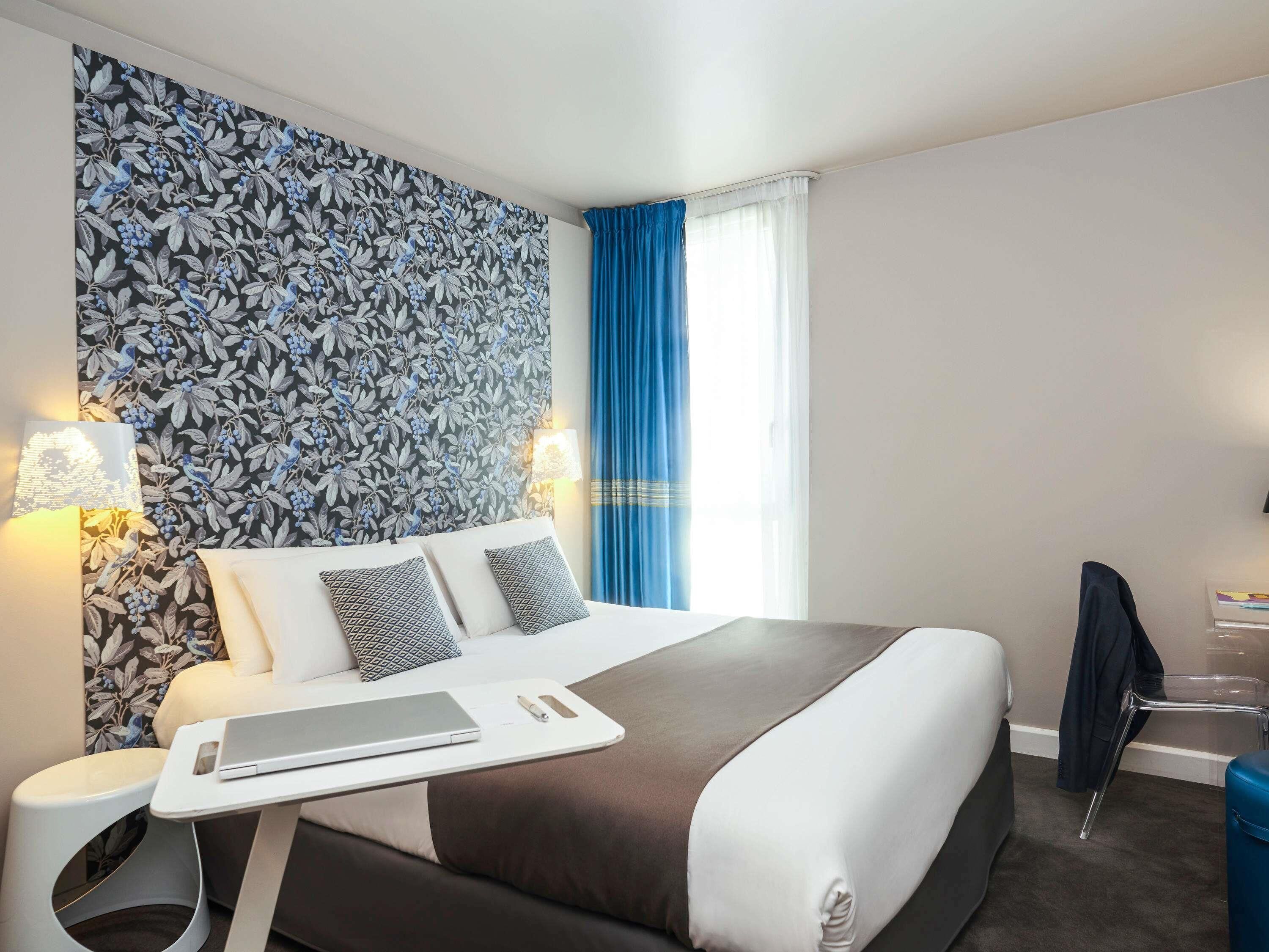 Habitación de hotel con una cama grande, dos lámparas de noche, una laptop sobre una mesita auxiliar, una cortina azul y una silla.