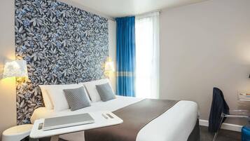 Habitación de hotel con una cama grande, dos lámparas de noche, una laptop sobre una mesita auxiliar, una cortina azul y una silla.