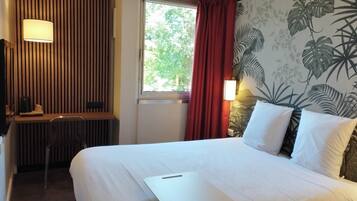 Habitación de hotel con cama, escritorio, silla, ventana con cortinas y un mural de plantas en la pared.