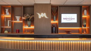 Un mostrador de recepción con acabado de madera, un gran logotipo iluminado con la letra 'M', una pantalla digital que muestra 'Mercure Hotels' y diversos objetos decorativos, como un jarrón con flores y una escultura.