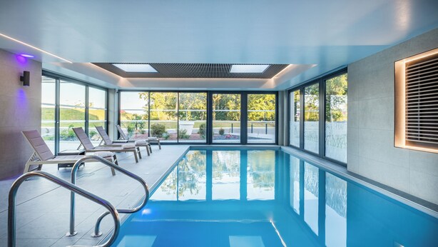 Una piscina interior moderna con paredes de vidrio transparente, techo iluminado por el sol y sillones de descanso a un lado.