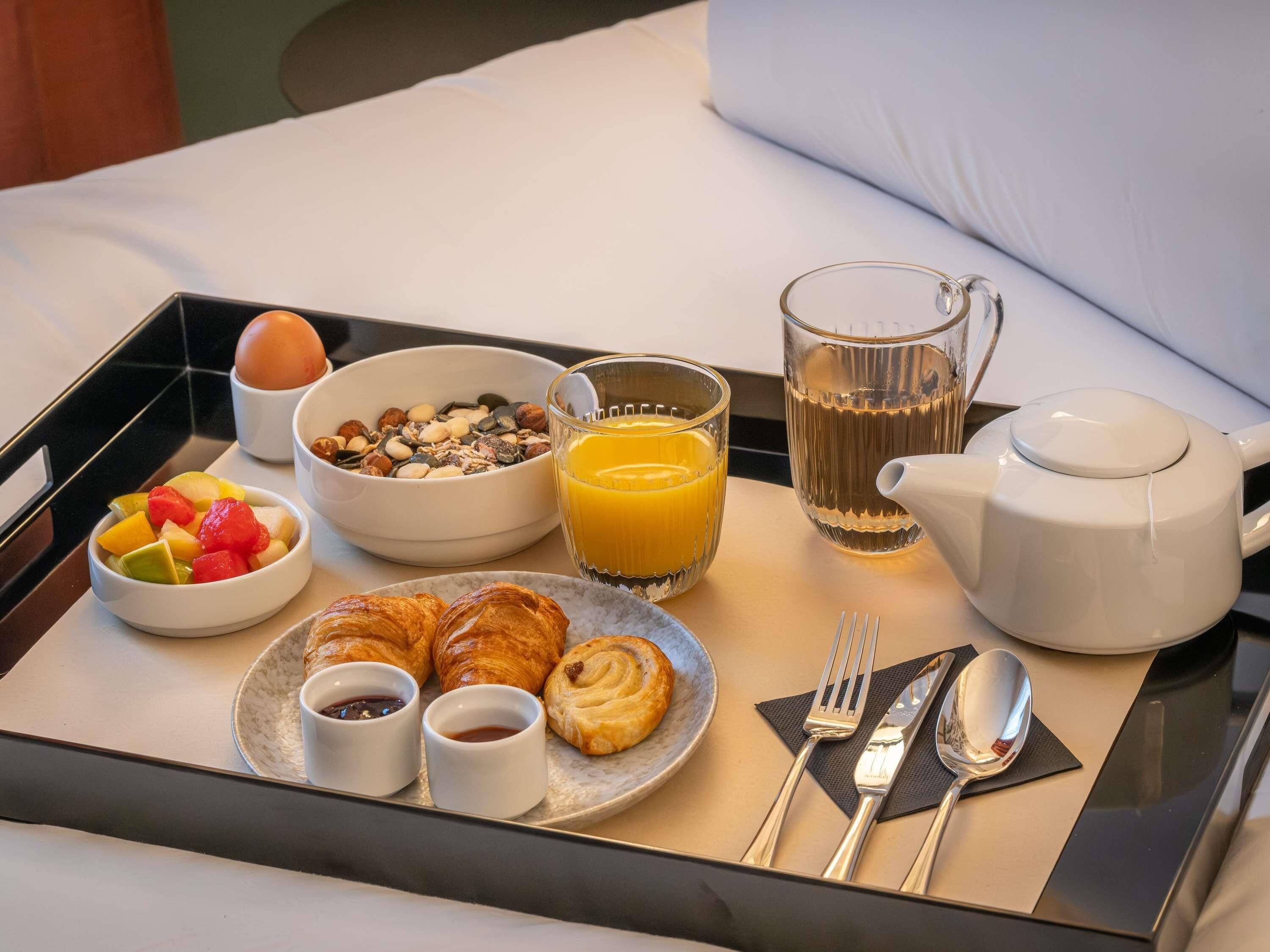 Bandeja de desayuno con cruasanes, fruta, granola, jugo de naranja y una tetera sobre la cama.