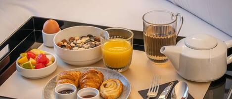 A breakfast tray with croissants, fruit, granola, orange juice, and a teapot on a bed.
