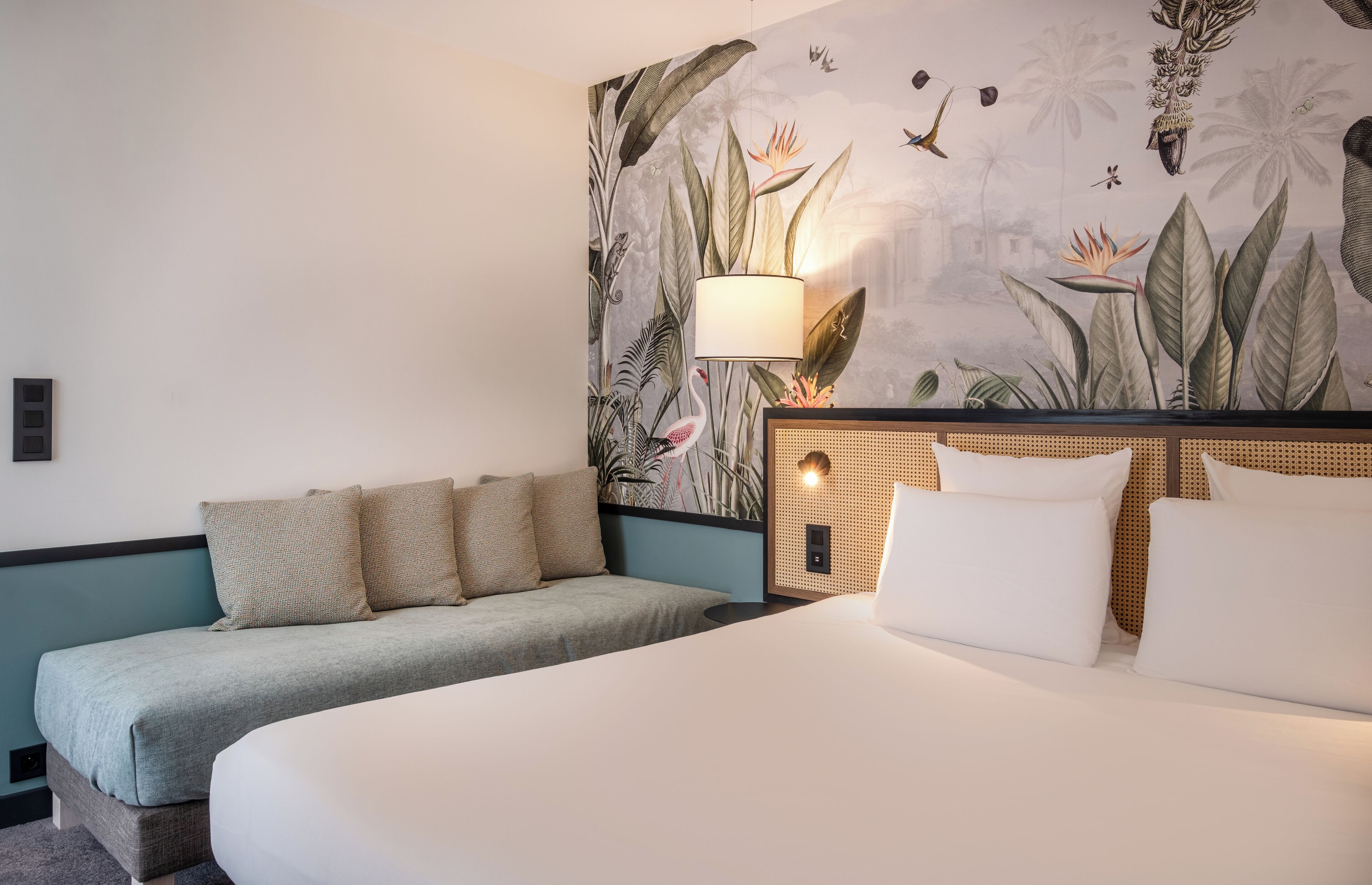 A hotel room with a bed, a sofa, a wall mural, a lamp, and a small table.