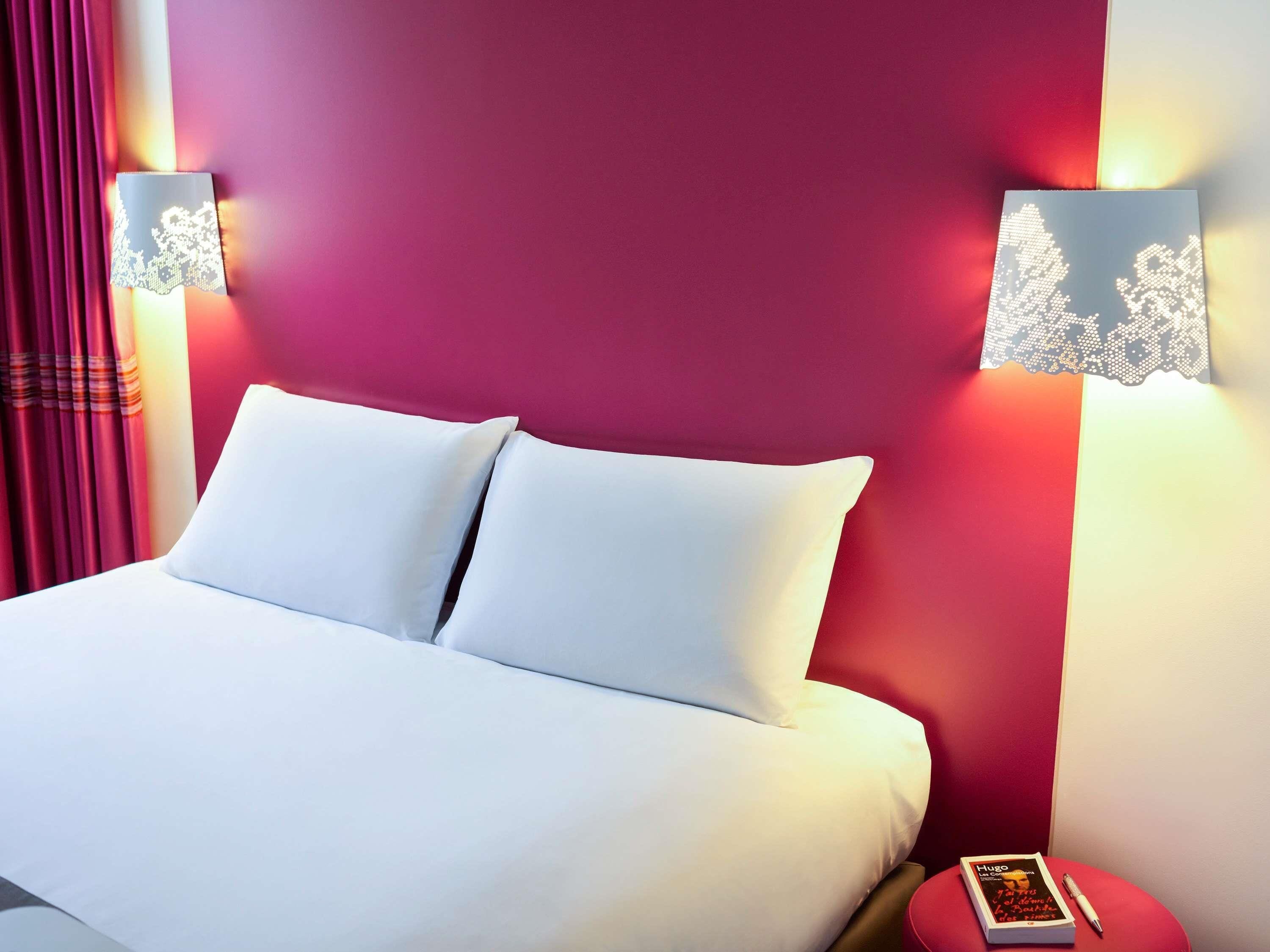 A hotel room with a bed, pillows, a nightstand, a lamp, and a book.