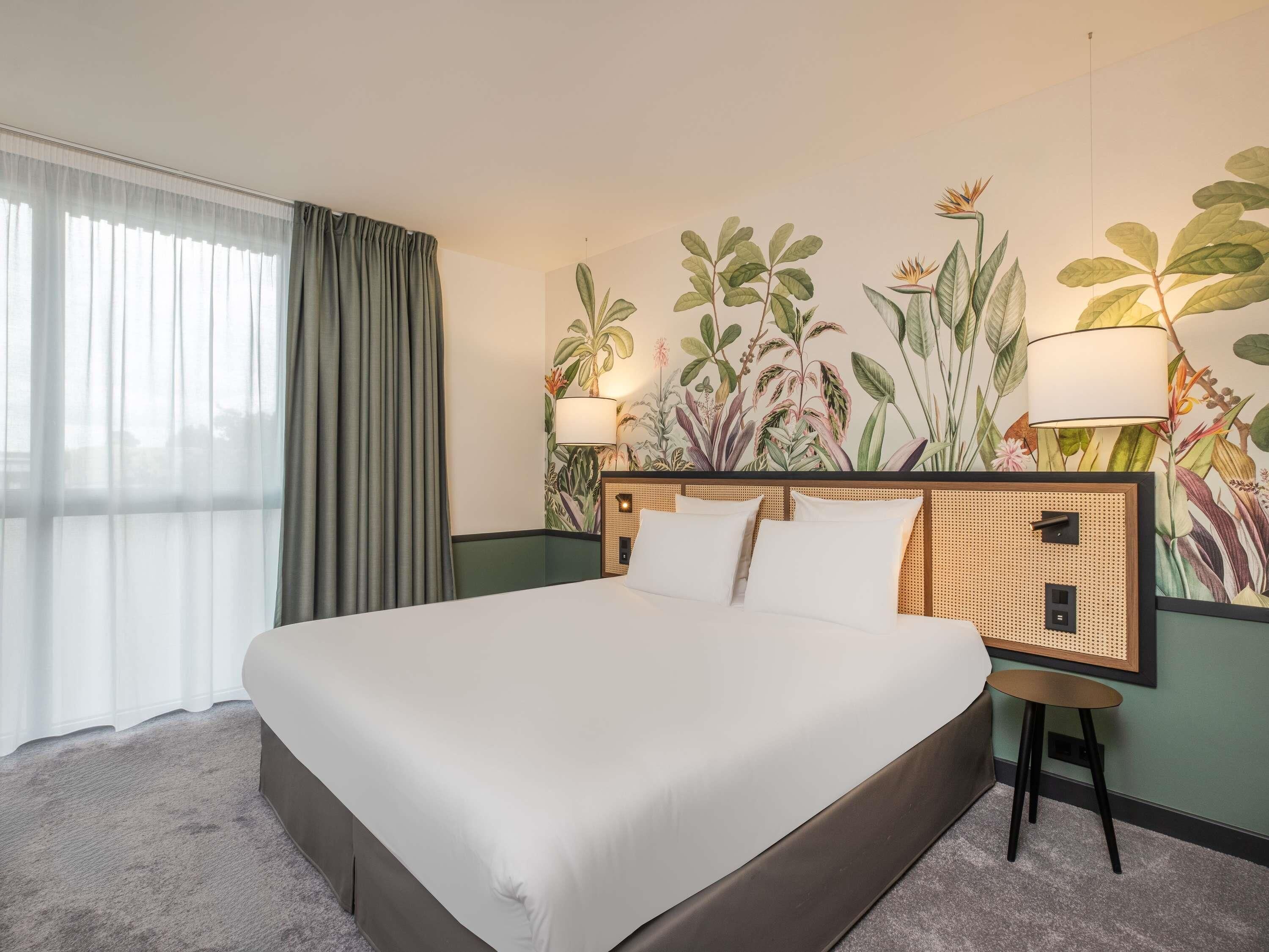 A hotel room with a large bed, a rattan headboard, a wall-mounted lamp, and a window with curtains.