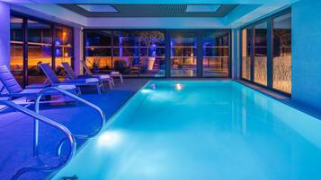 A modern indoor pool with clear glass walls, lounge chairs, and a view of the night sky.