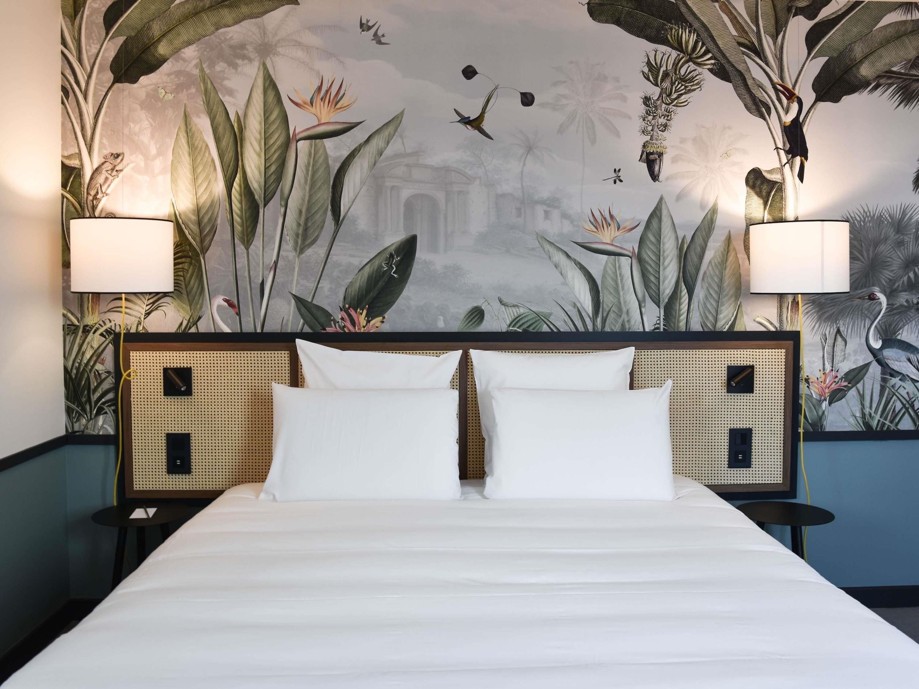 A bed with white linens, two bedside lamps, and a wall mural with tropical plants and birds.