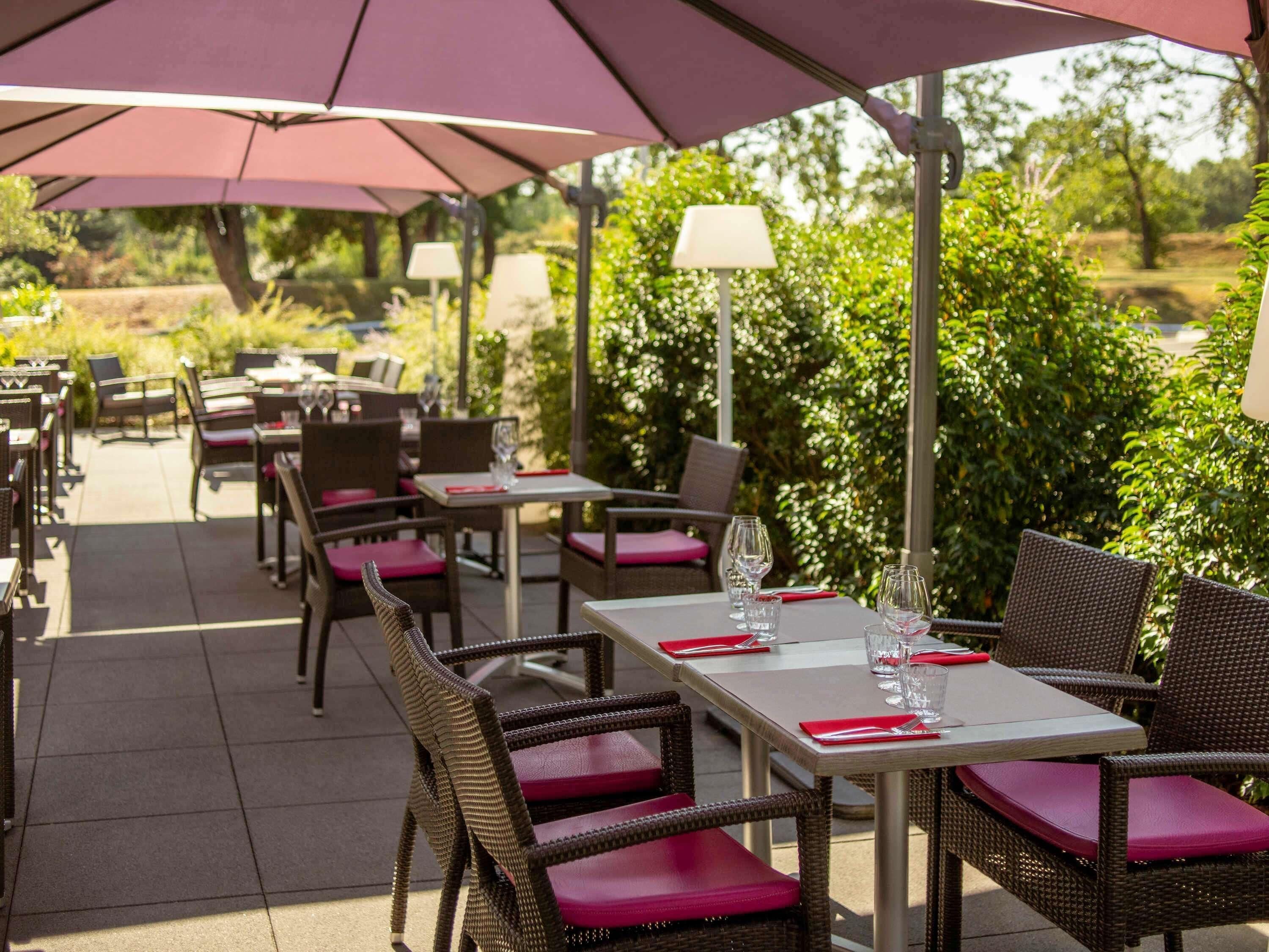 Un área de comedor al aire libre con sillas y mesas de mimbre, cubierta por grandes sombrillas y rodeada de vegetación.