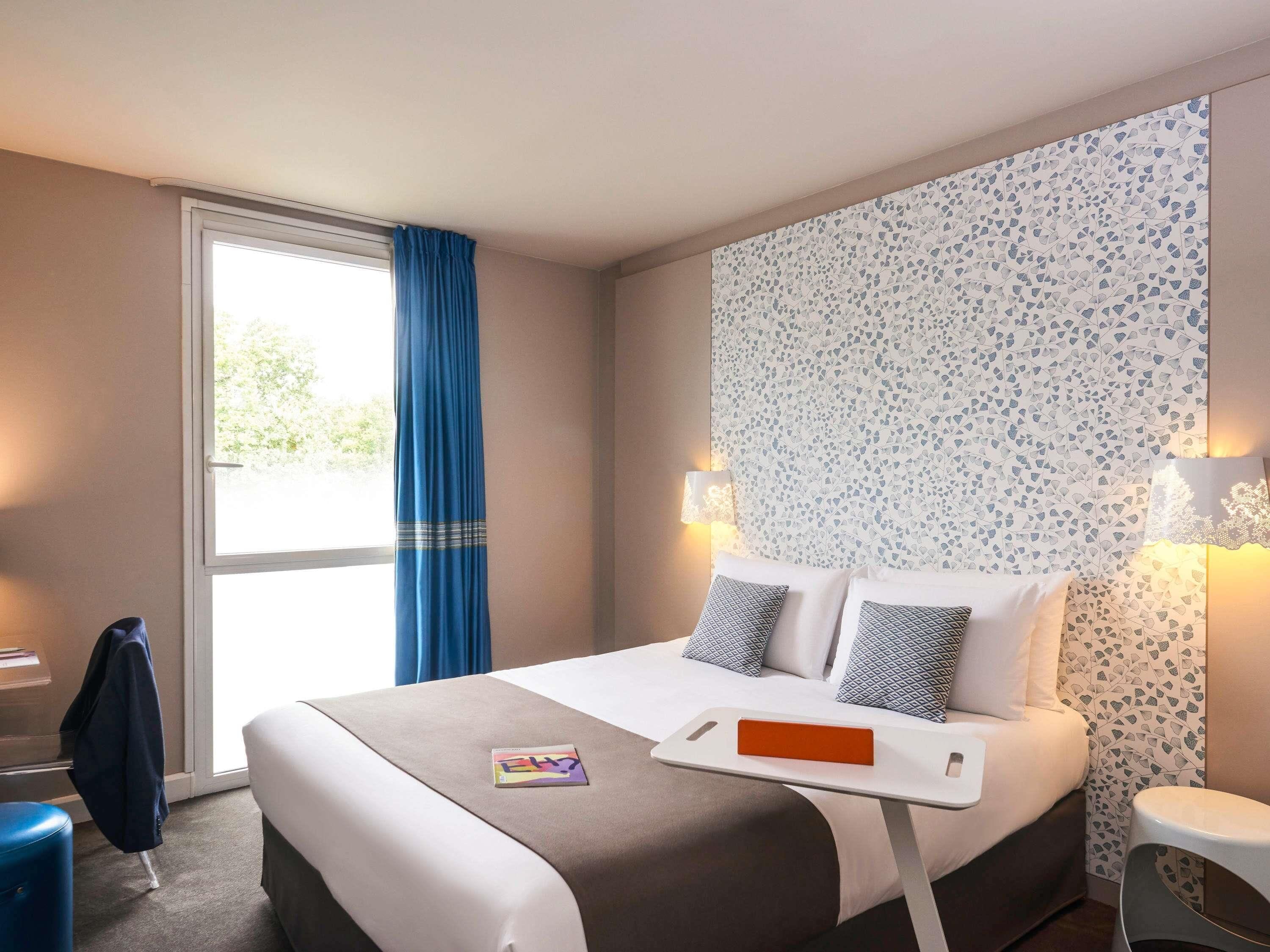 Una habitación de hotel con una cama, un escritorio, una silla, un ventanal con cortinas y una pared decorada con un diseño de hojas.