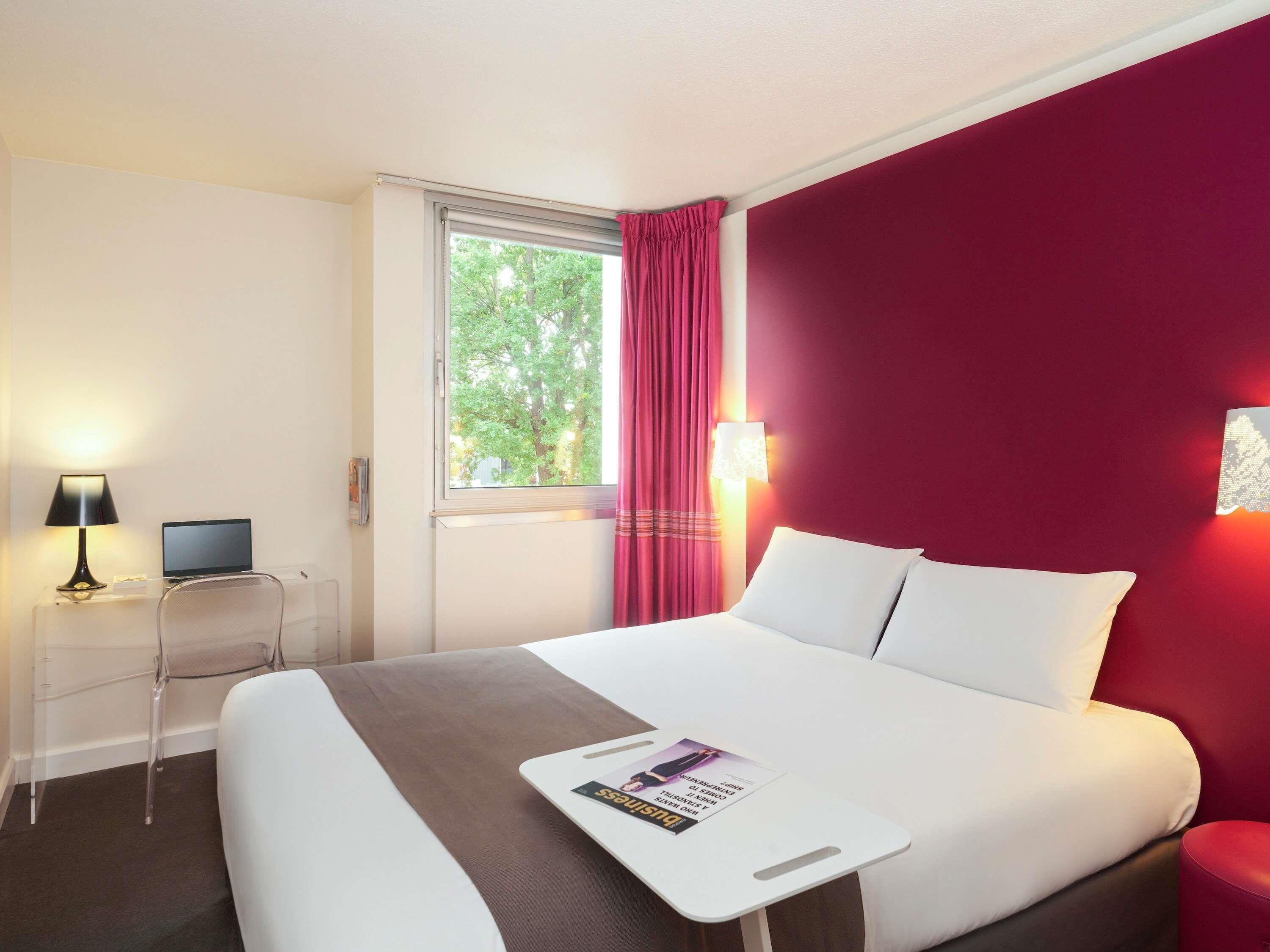 Habitación de hotel con cama, escritorio con computadora portátil, ventana con cortinas y pared roja.