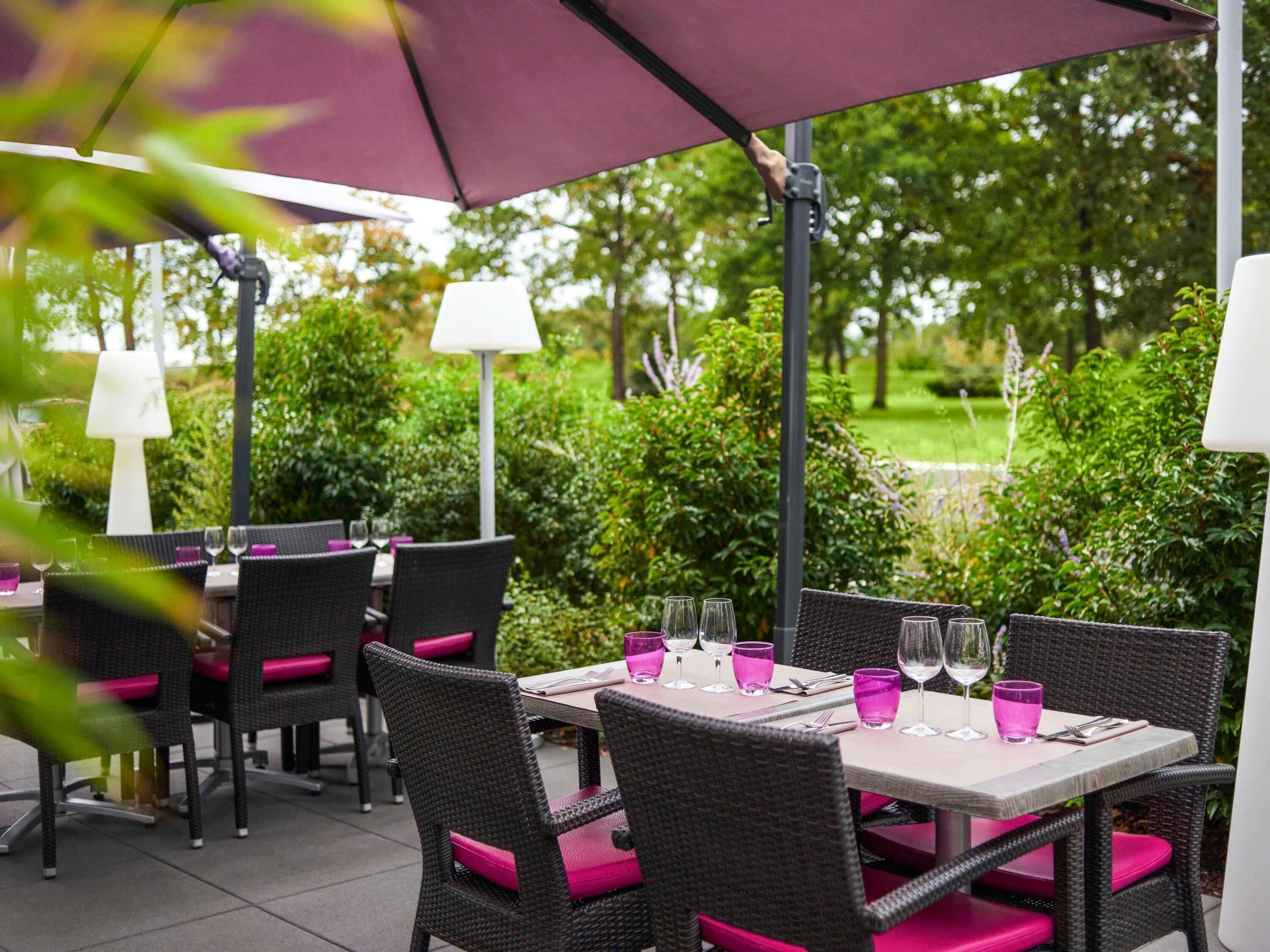 Un espace de restauration en plein air avec des chaises et des tables en osier, chaque ensemble étant agrémenté d’une nappe blanche et de coussins roses.