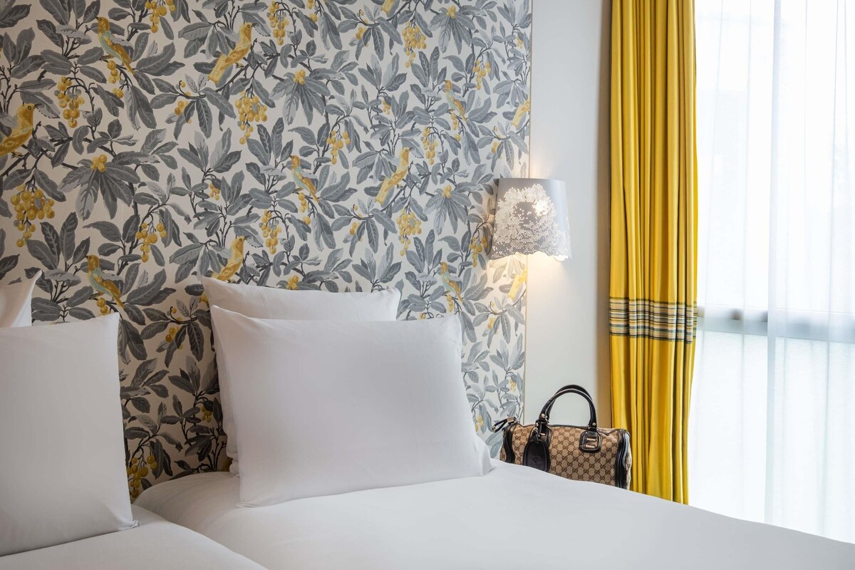 A hotel room with a floral wallpaper, a bed with white bedding, a lamp, and a yellow curtain.