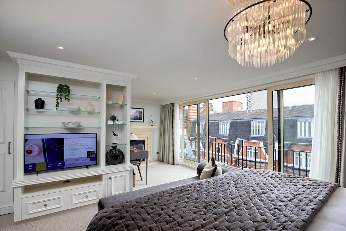Een slaapkamer met een groot bed, een televisie op een witte kast, een kroonluchter en uitzicht op gebouwen door grote ramen.