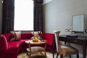 A modern hotel room with a red sofa, a wooden dining table with fruits, a bottle of wine, and a desk with a mirror. - The Capital Hotel, Apartments & Townhouse - Small Luxury Hotels of The World (London)