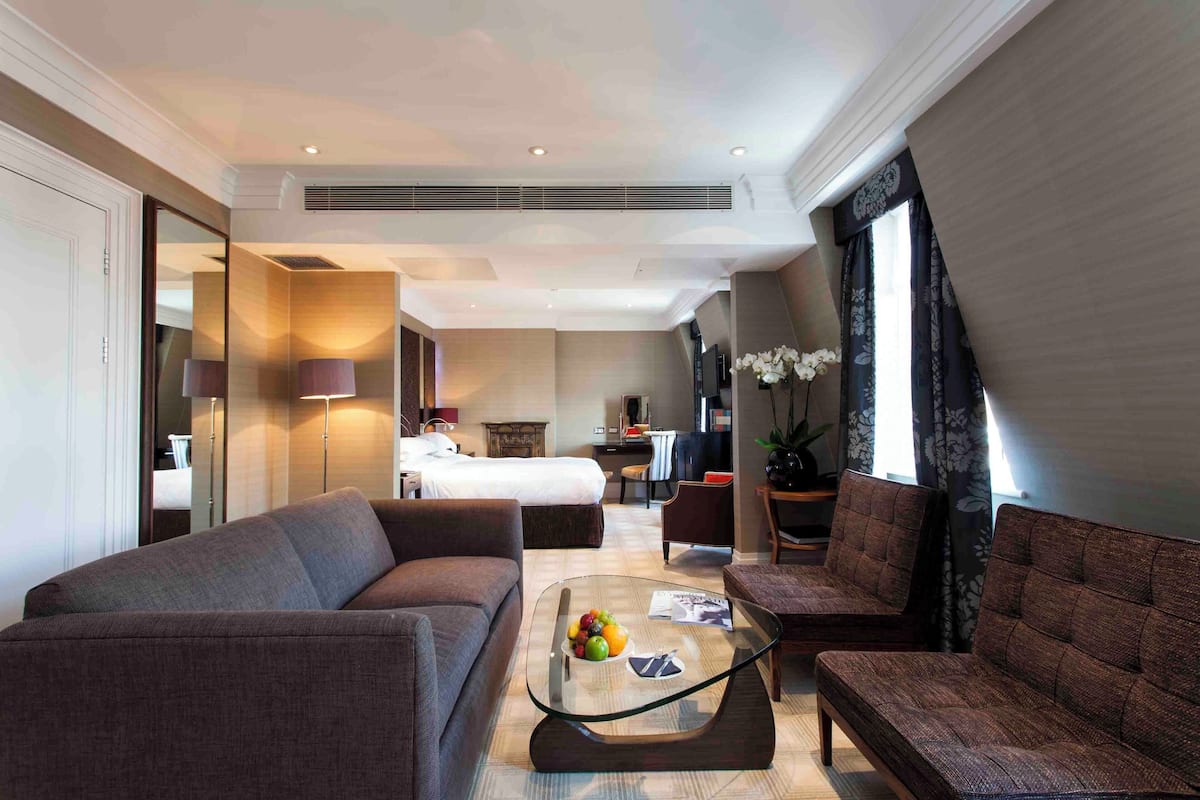 Een moderne hotelkamer met een bank, fauteuil en een glazen bijzettafeltje met fruit. Achtergrond is een bed en een groot raam met gordijnen.