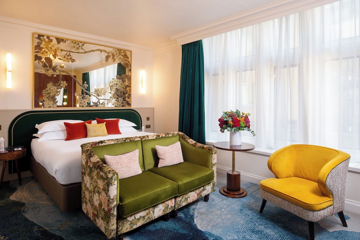 Een hotelkamer met een bed, een groene bloemenbank, een gele fauteuil, een ronde tafel met een bloevas en een grote spiegel aan de muur.