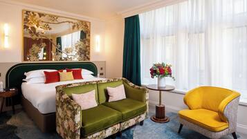 Et hotelværelse med en seng, en grøn blomstrende sofa, en gul lænestol, et rundt bord med en vase blomster og et stort spejl på væggen.