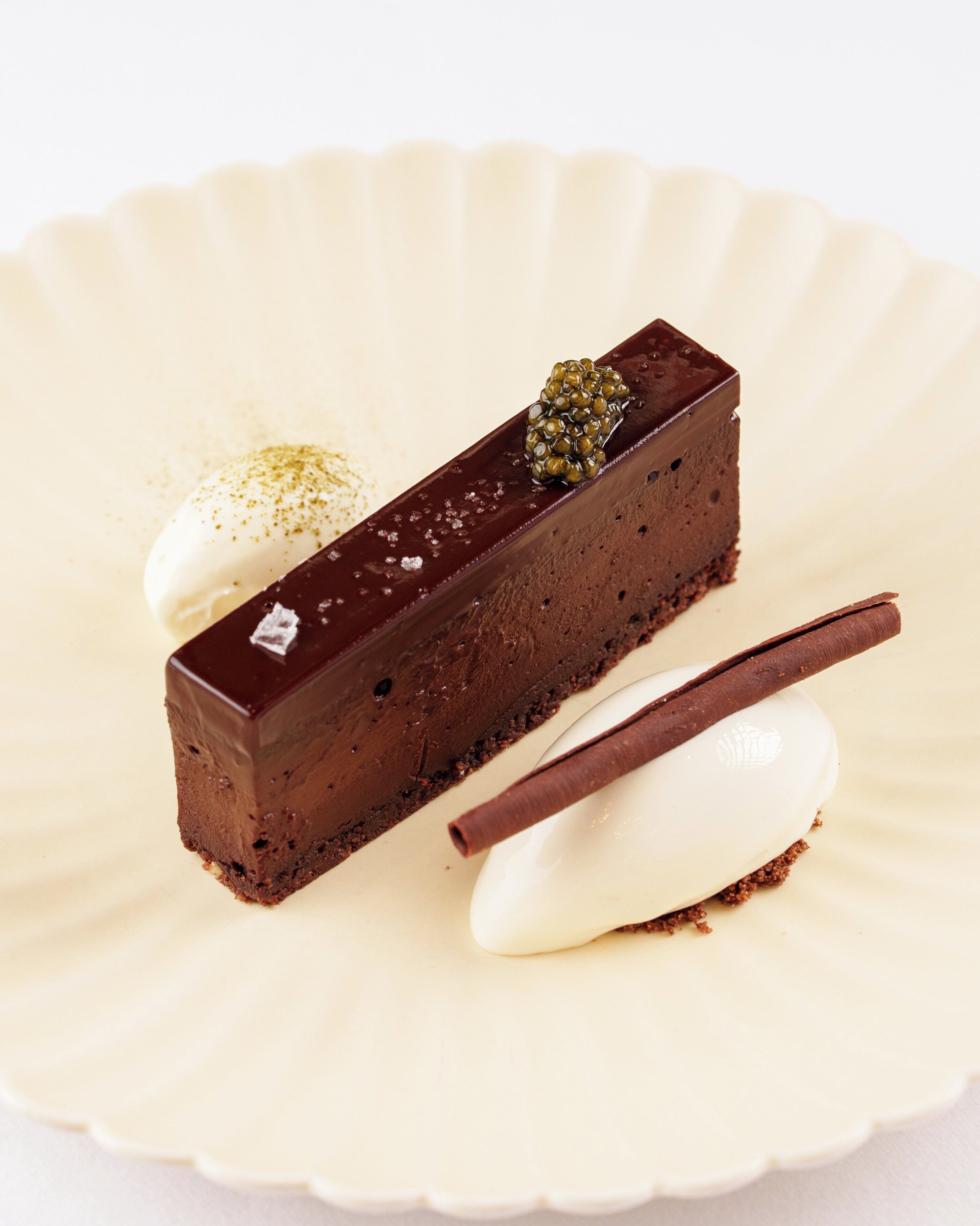 A slice of chocolate cake with a glossy chocolate glaze and a decorative chocolate piece on top, served on a white plate with a scalloped edge.