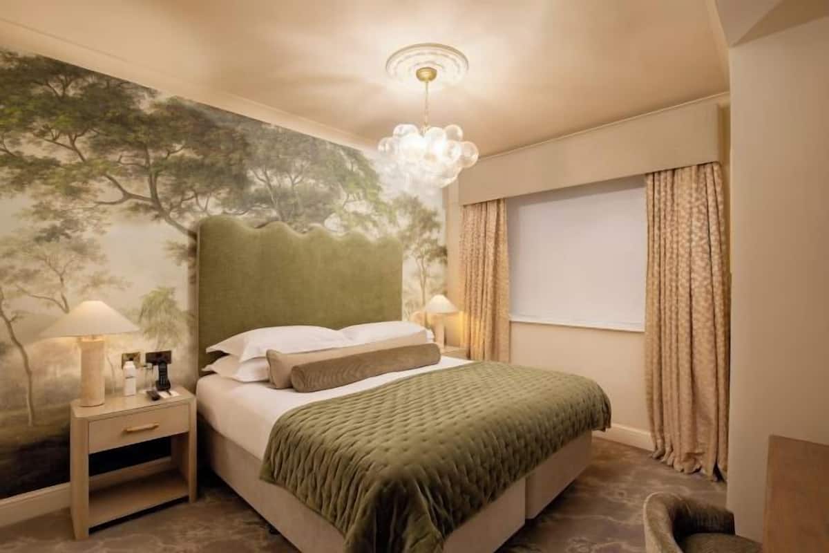 Een slaapkamer met een bed, nachtkastjes, een muurschildering, een raam met gordijnen en een kroonluchter.
