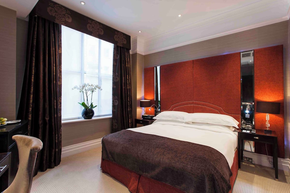Een hotelkamer met een groot bed, donkere gordijnen, een vaas met bloemen, een bureau en een nachtkastje met een lamp.